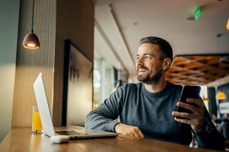 Mann sitzt lächelnd mit Smartphone und Laptop in einem Café.