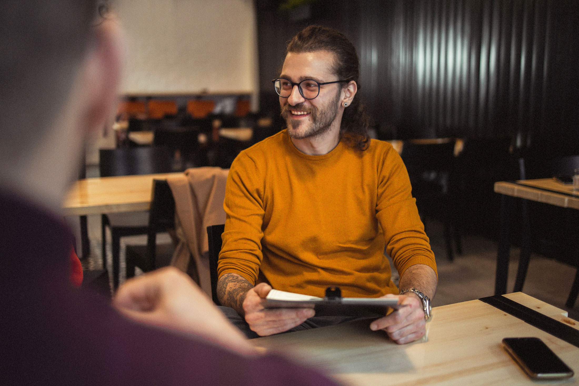 Mann sitzt an einem Tisch, hält ein Tablet und lächelt seinen Gesprächspartner an.